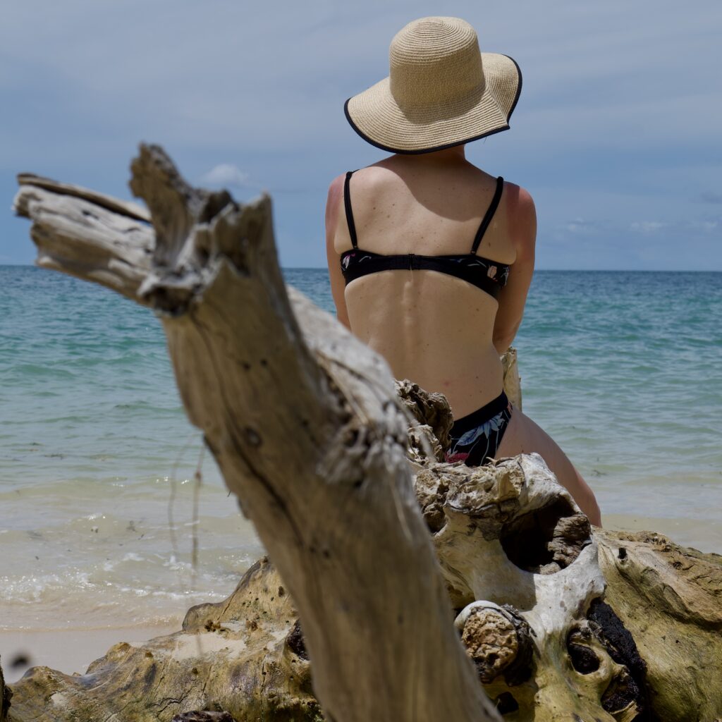 Femme de dos en maillot de bain et chapeau de paile, assise sur des rochers face à l'océan à Sumba.