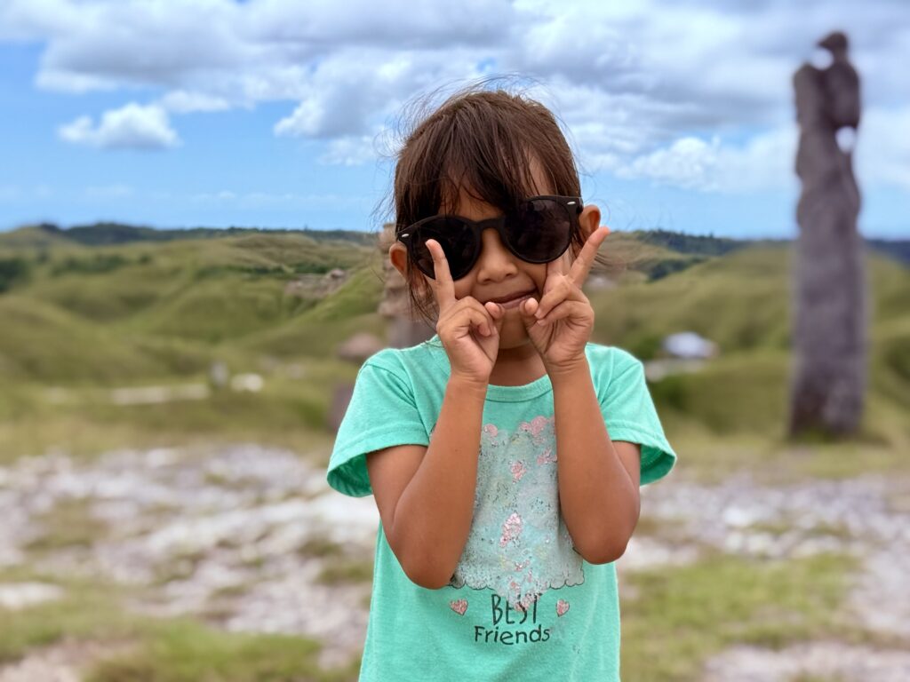 Petite fille de Sumba souriante, portantr des lunettes de solei et faisant le signe de la paix avec les doigts.