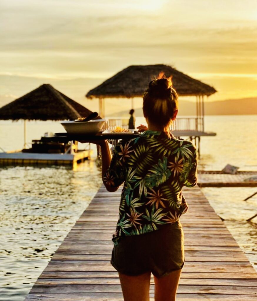 Une femme portant un plateau de plats marche sur un ponton en bois vers des huttes sur pilotis, baignées par la lumière dorée du coucher de soleil à Sumba.